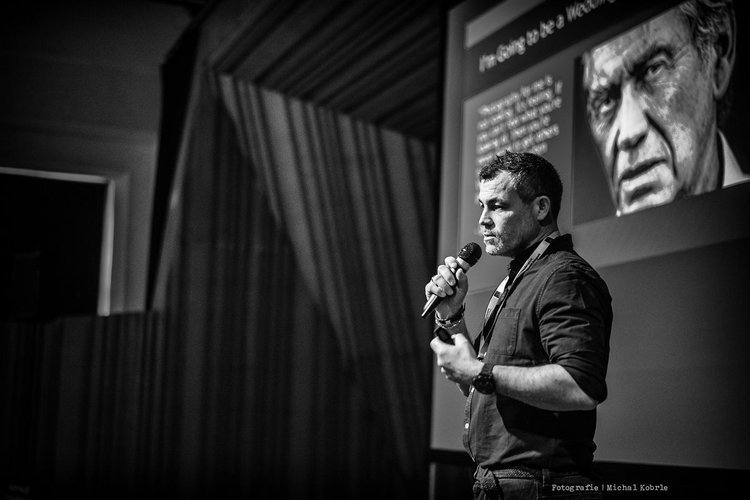 A man standing on stage holding a microphone, giving a presentation with a large screen behind him displaying a facial portrait and text.