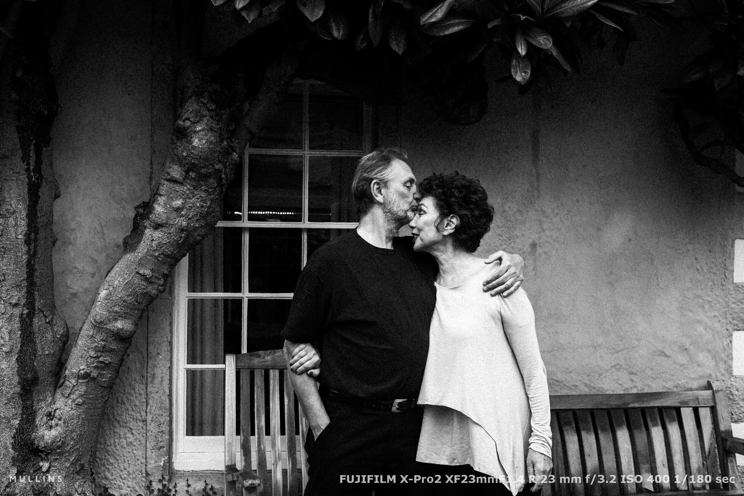 Older couple sharing a kiss outside their cottage beneath a tree, a moment of affection captured naturally
