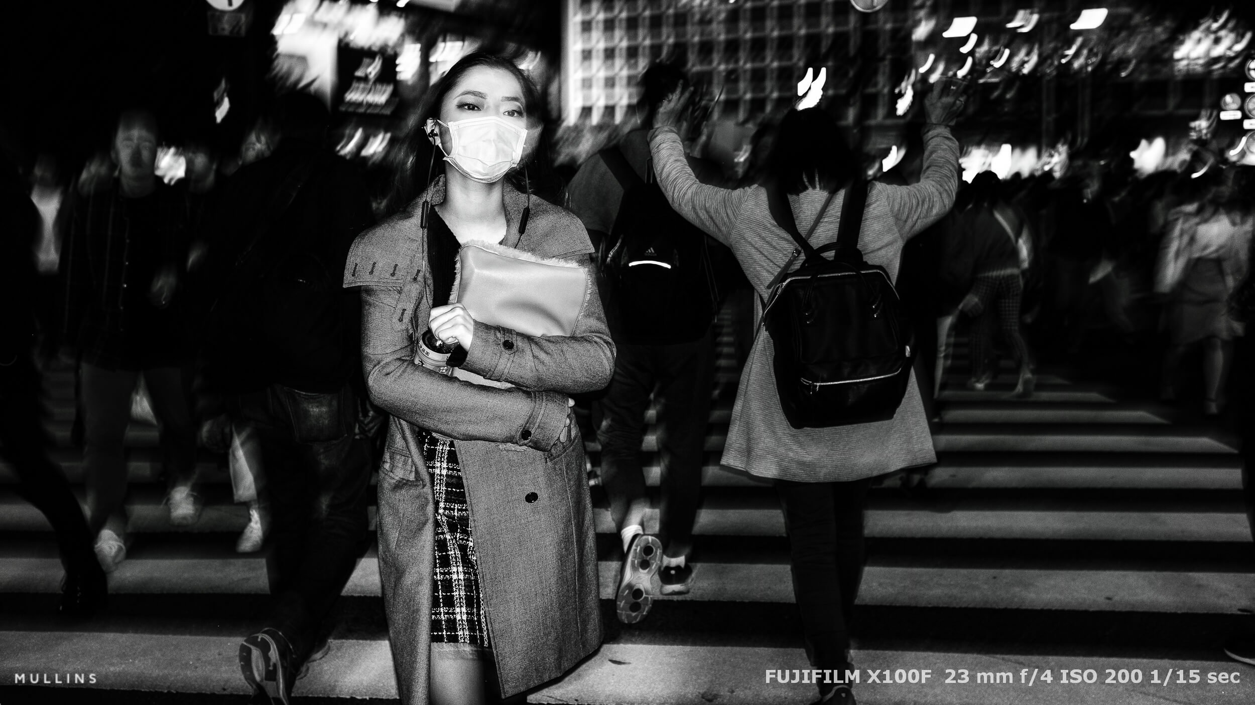 Masked woman crossing a street at night, surrounded by blur and city lights. She holds a bag and wears earphones.