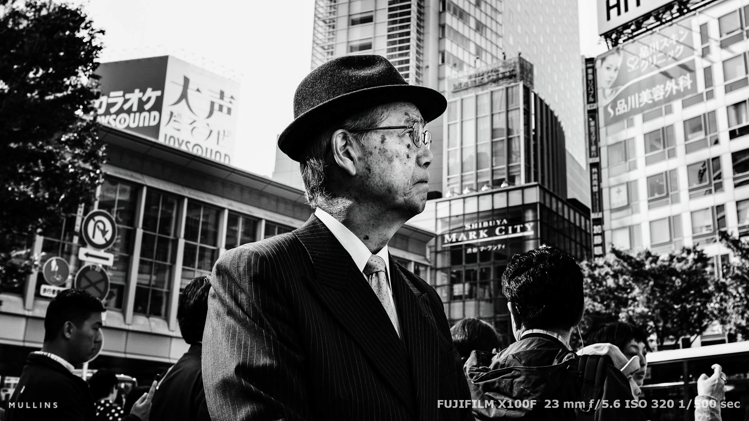 Elderly man in bowler hat stands on busy Tokyo street corner. Black and white shot with signage and sharp contrast.