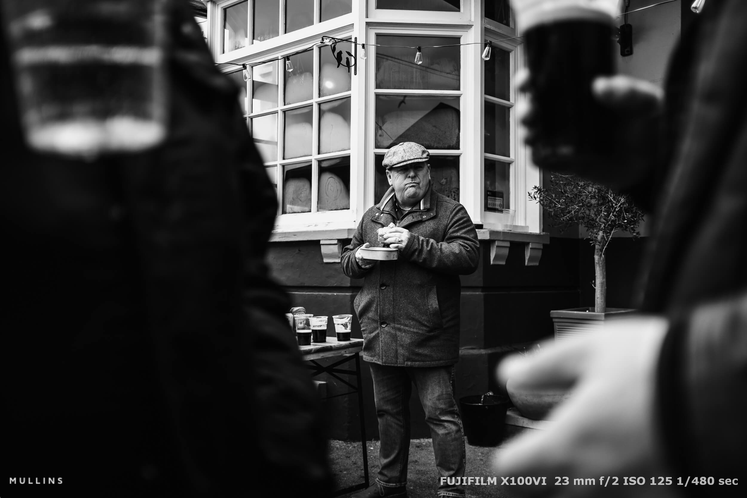 Man eating a burger outside a pub, framed between two pints of Guinness. Candid street moment with layered focus.
