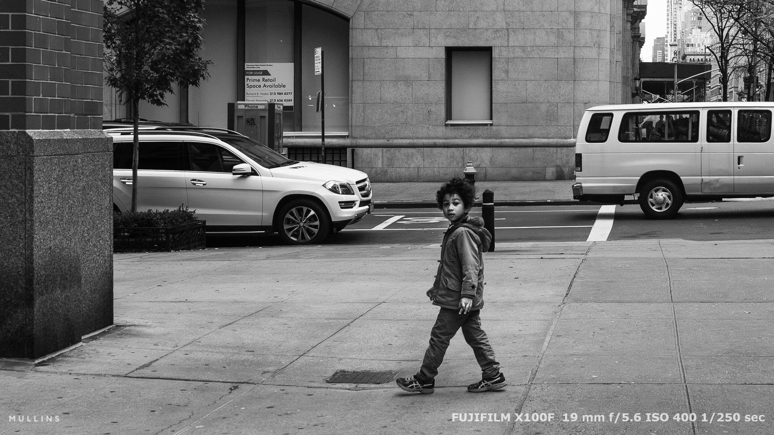 Boy with curly hair walking across NYC street, looking back. Black and white photo with cars and soft light in the background.