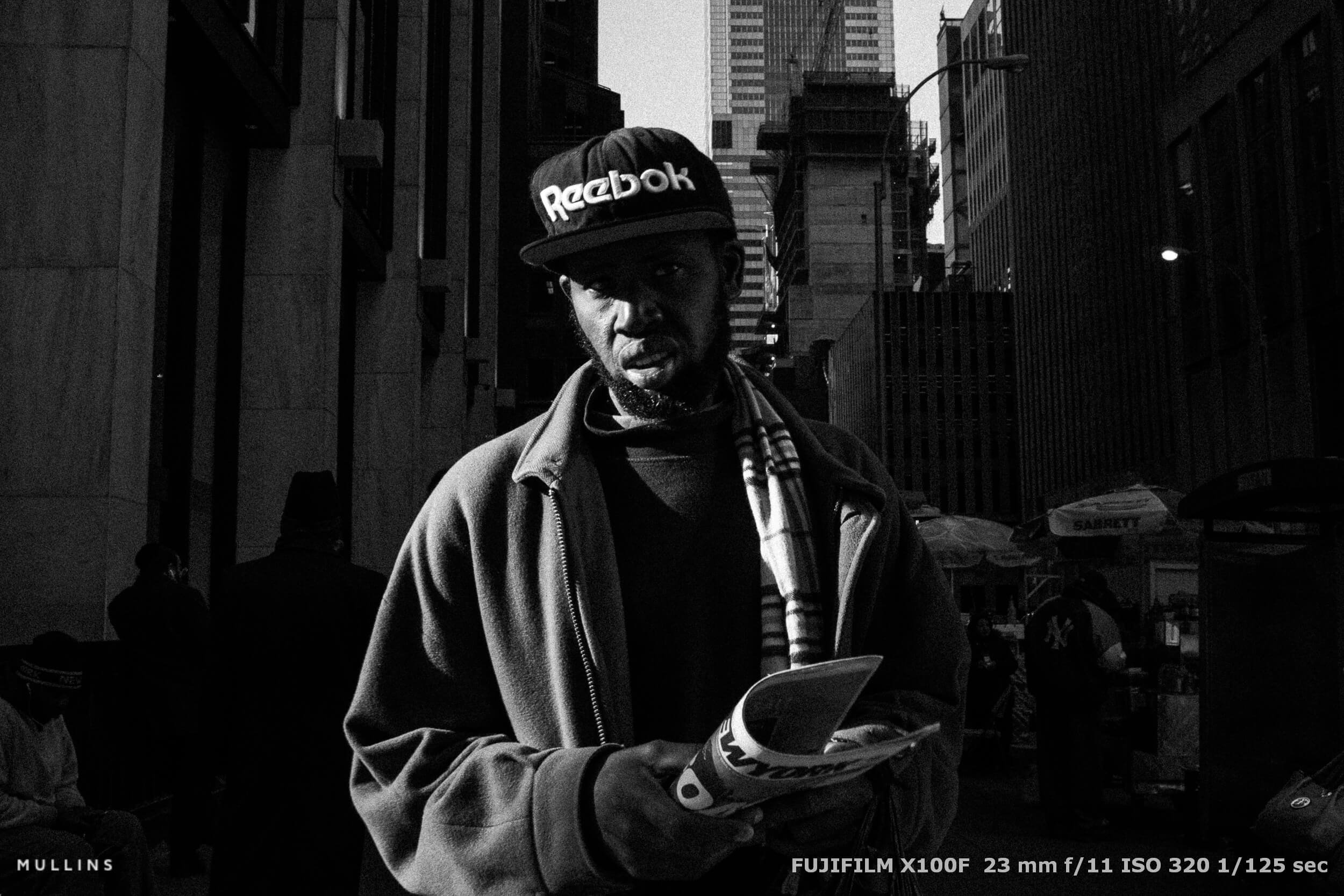 Man in Reebok cap holding newspaper, lit by strong sunlight. High-contrast black and white city scene with tall buildings behind.