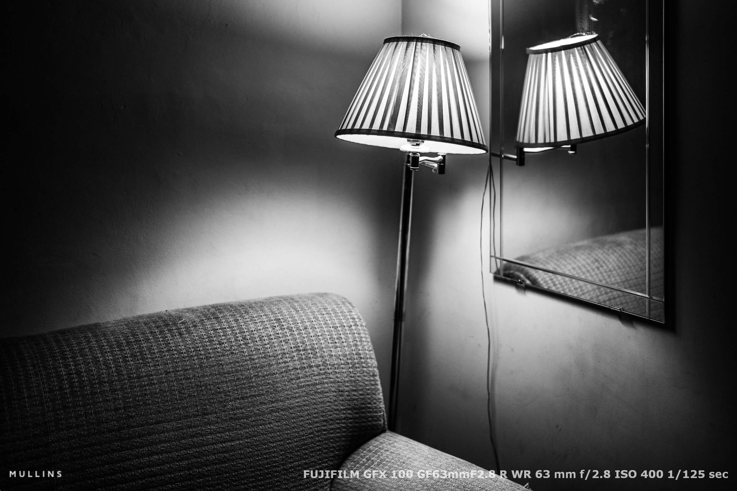 Still life of a striped lampshade reflected in a mirror beside a textured armchair, lit with natural shadow and contrast