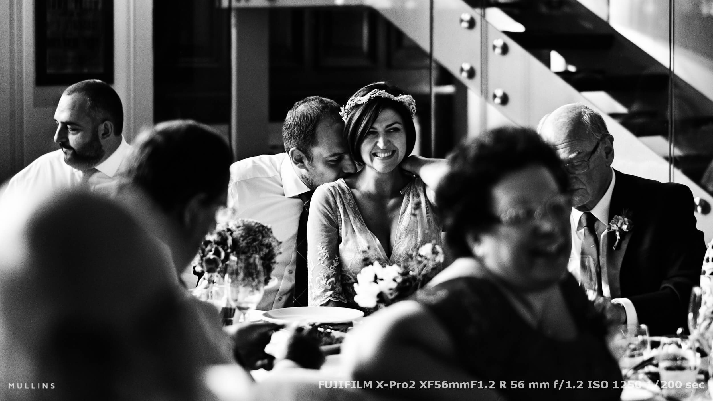 Newlyweds sharing a quiet laugh during their wedding breakfast, surrounded by guests in a relaxed and candid reception scene