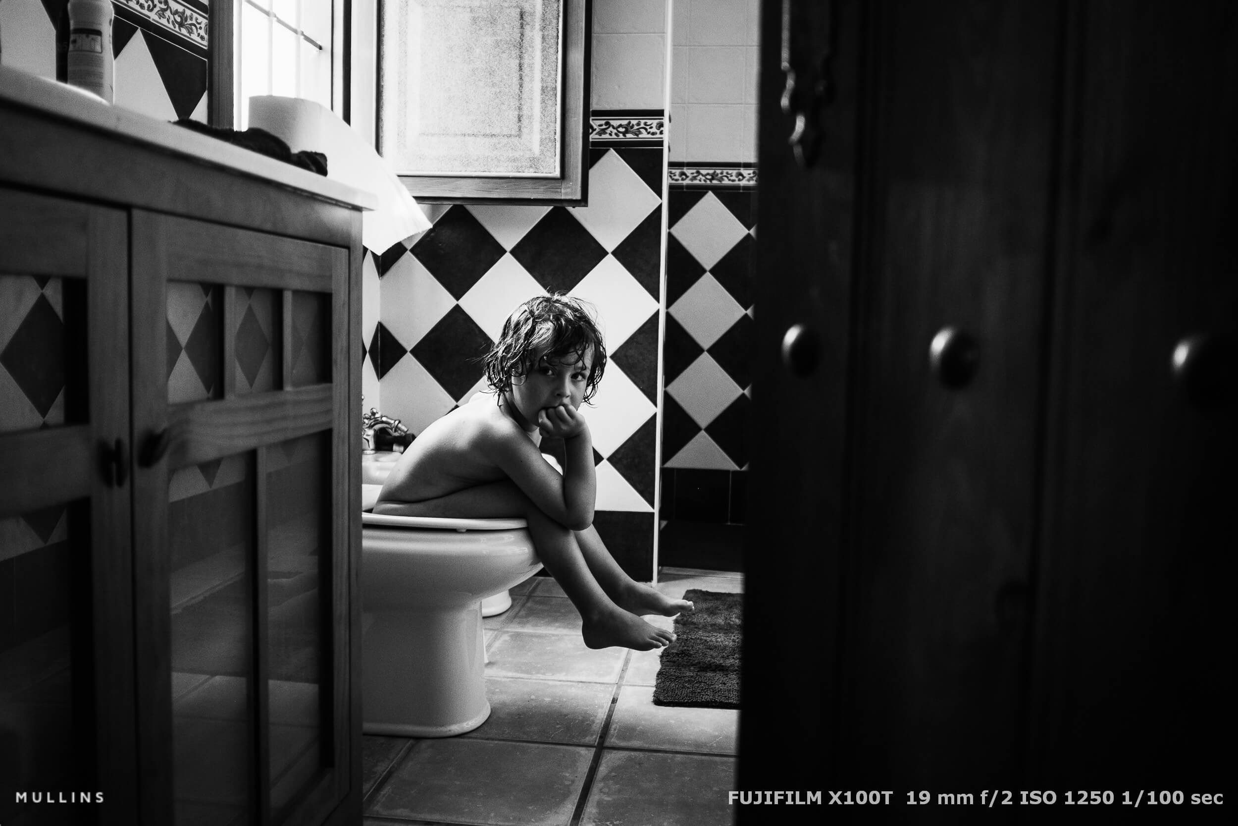 Unposed moment of a young child sitting on a toilet in a tiled bathroom, captured quietly through a doorway