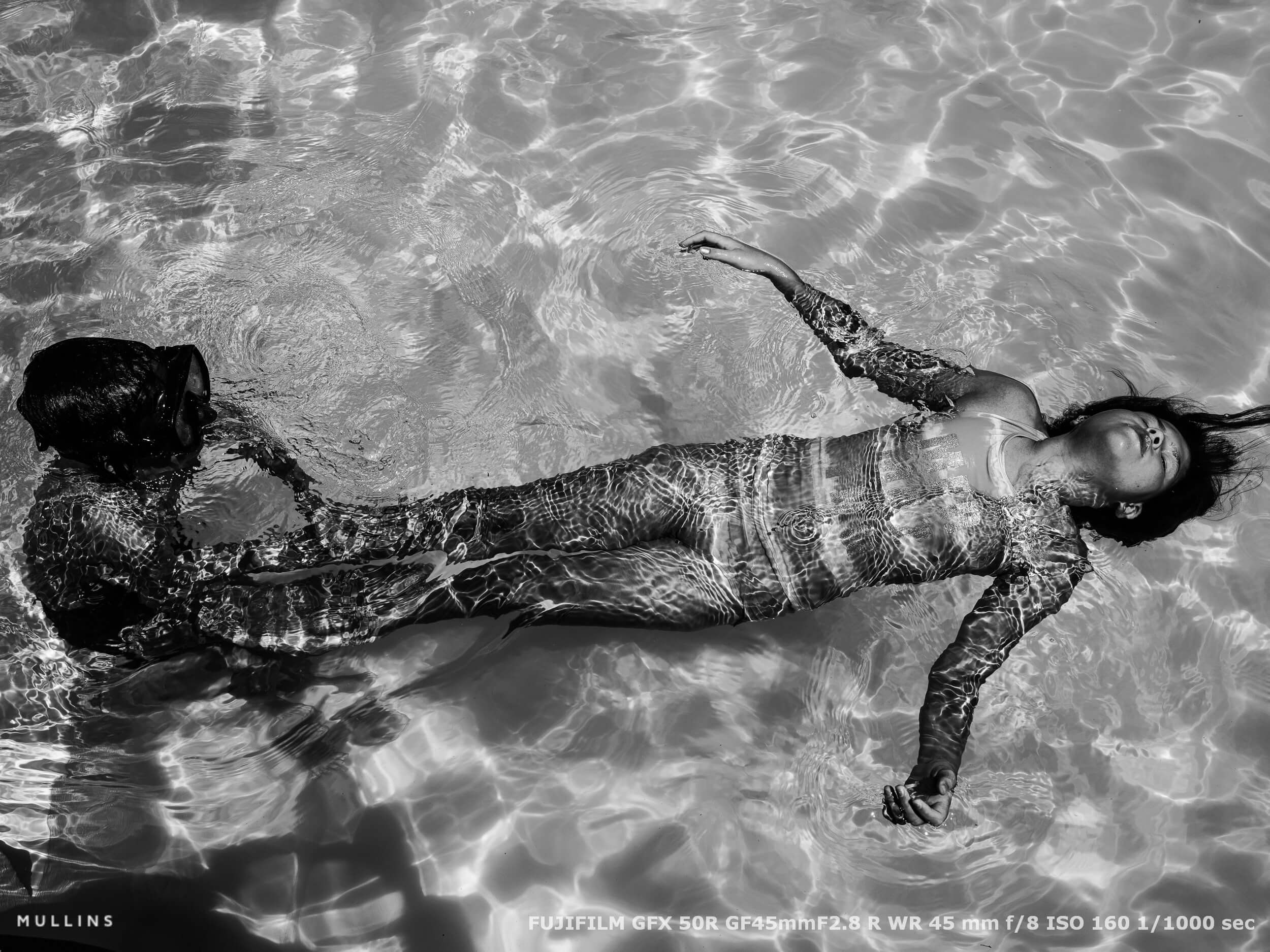 Child floating on her back in a sunlit swimming pool while another child swims nearby, captured with reflections and soft tones