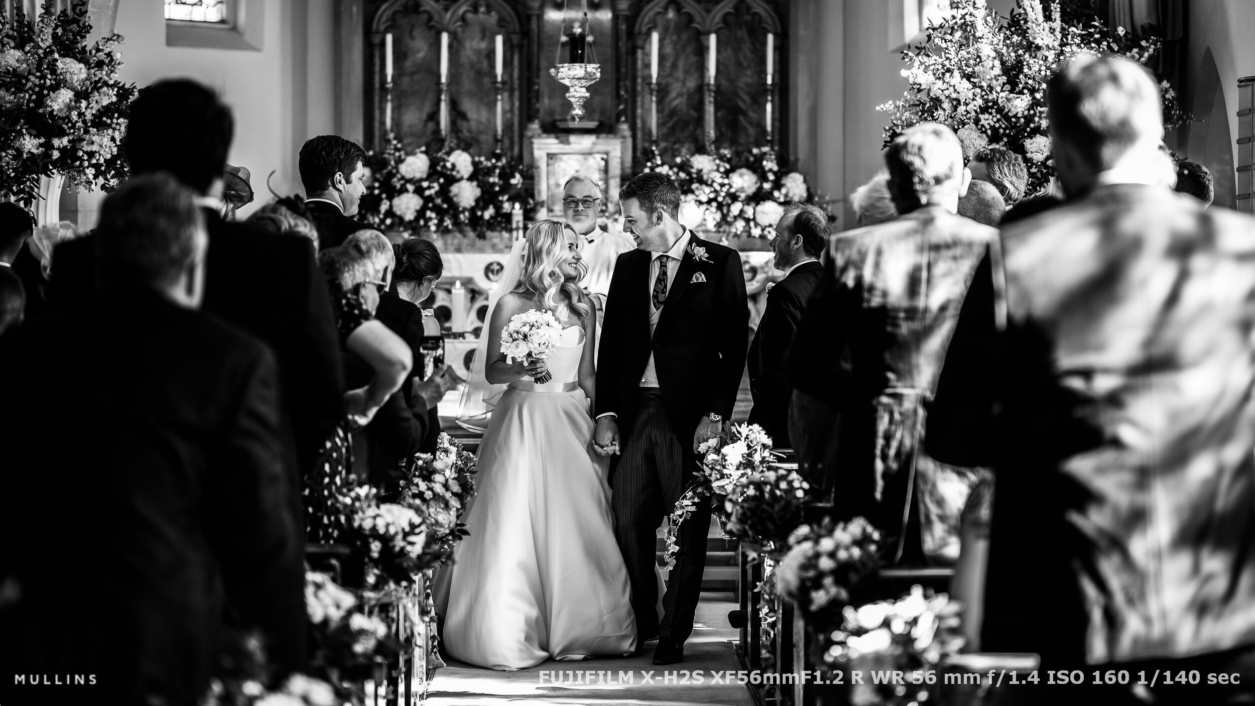 Bride and groom walking down the aisle hand in hand, smiling at each other just after their wedding ceremony, surrounded by guests