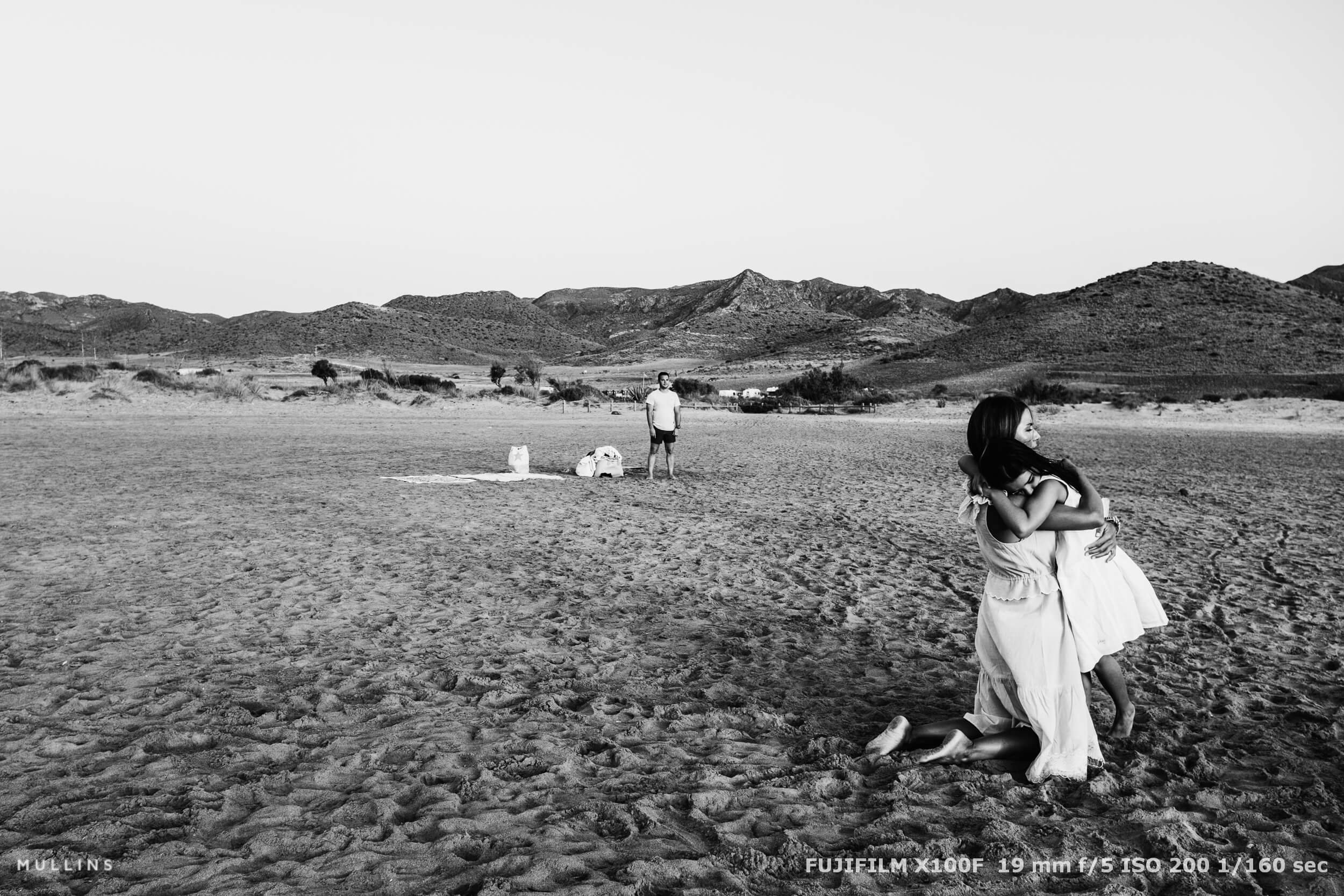 Mother and child embracing on a wide sandy beach in Spain while another family member stands in the background, captured in an unposed documentary style