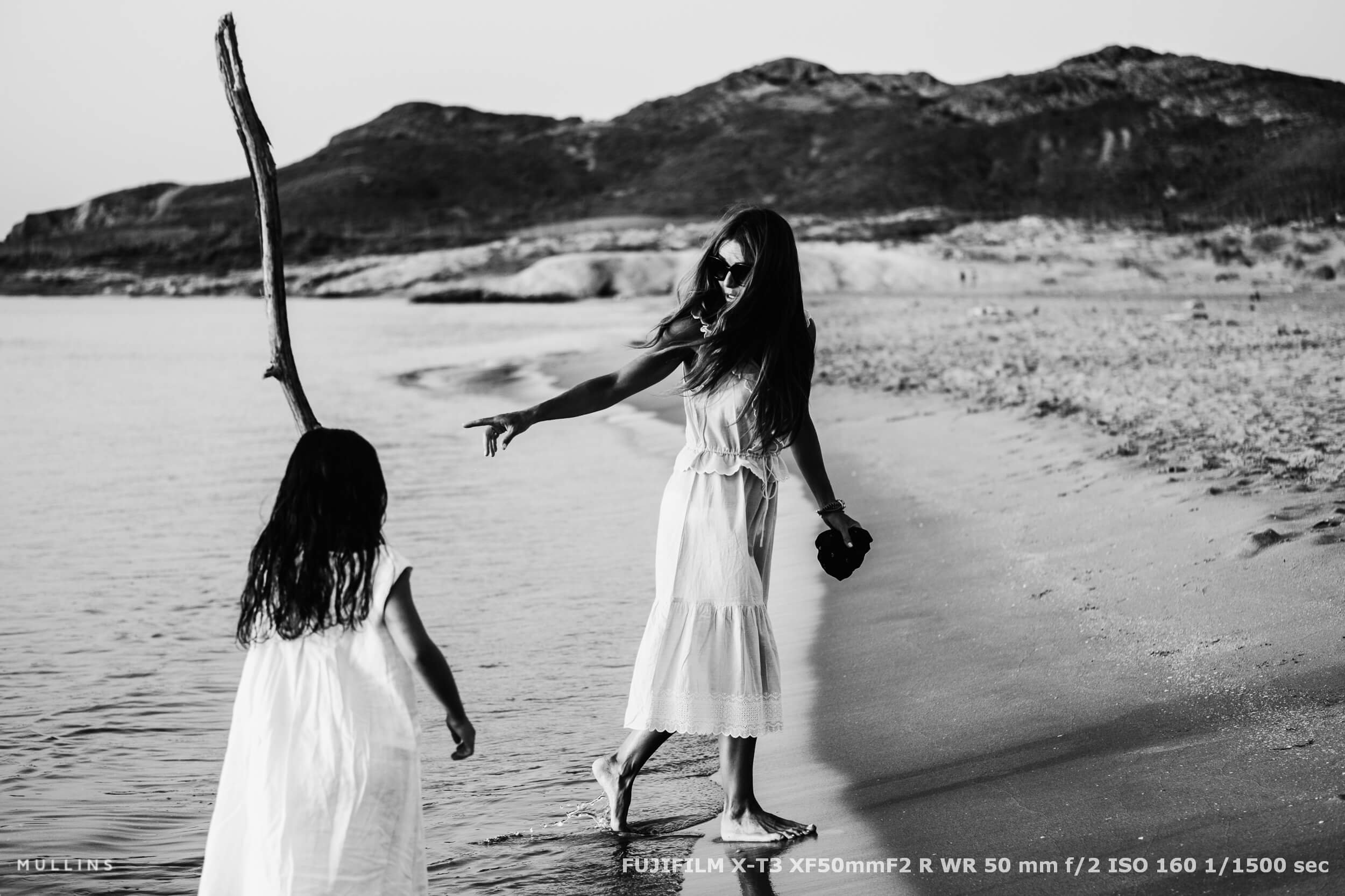 Mother and daughter walking along a quiet beach shoreline, the mother gesturing gently while barefoot in the water