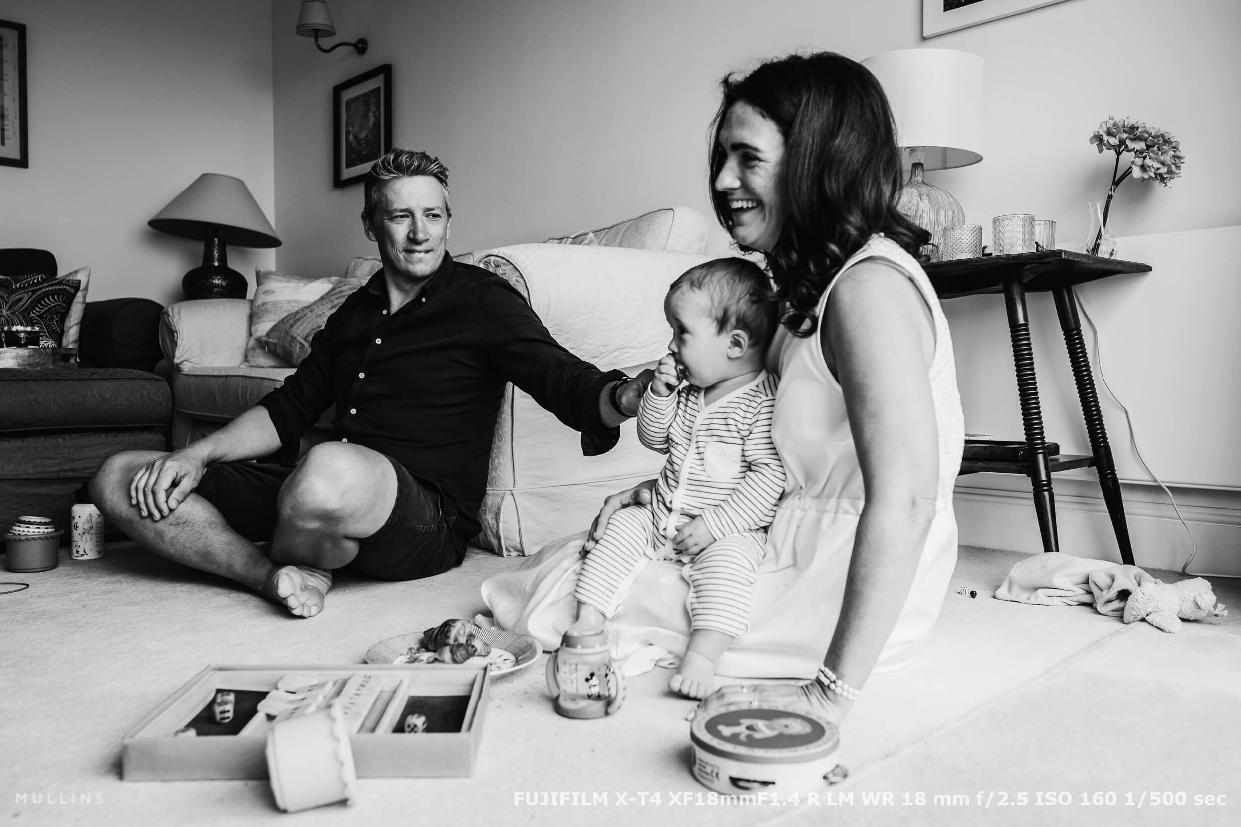Relaxed family moment in a living room, with parents and baby sitting on the floor surrounded by toys and soft light