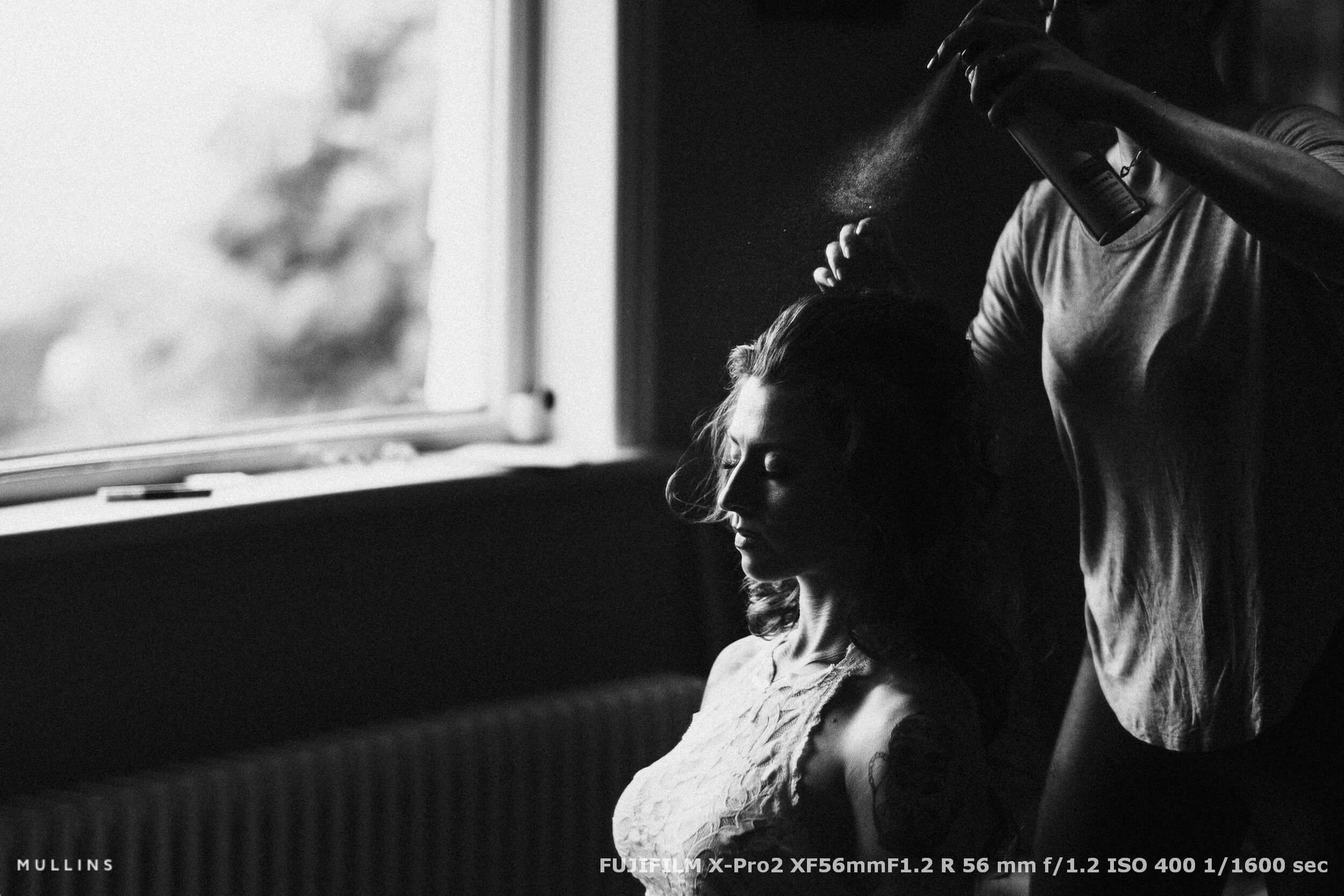 Bride in soft natural light as a stylist sprays her hair, caught mid-moment during the quiet preparations on the wedding morning