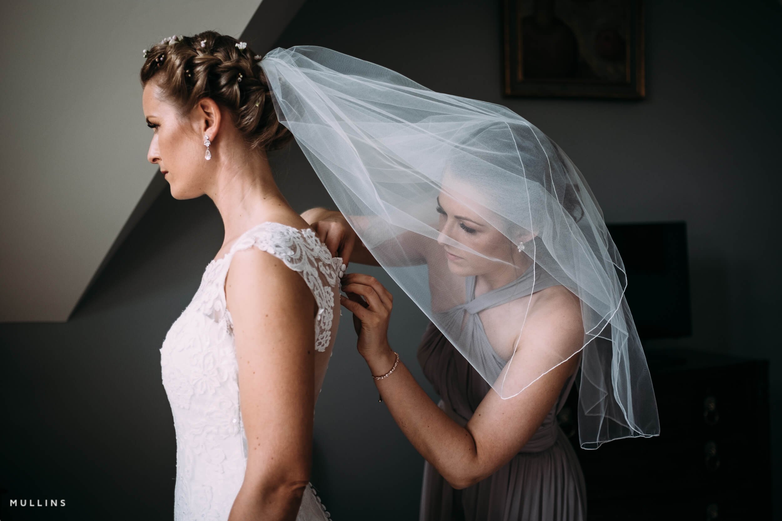 Bridal party preparing for a wedding, with a bride in a lace wedding dress and veil, and a bridesmaid assisting her with her dress in a dimly lit room.