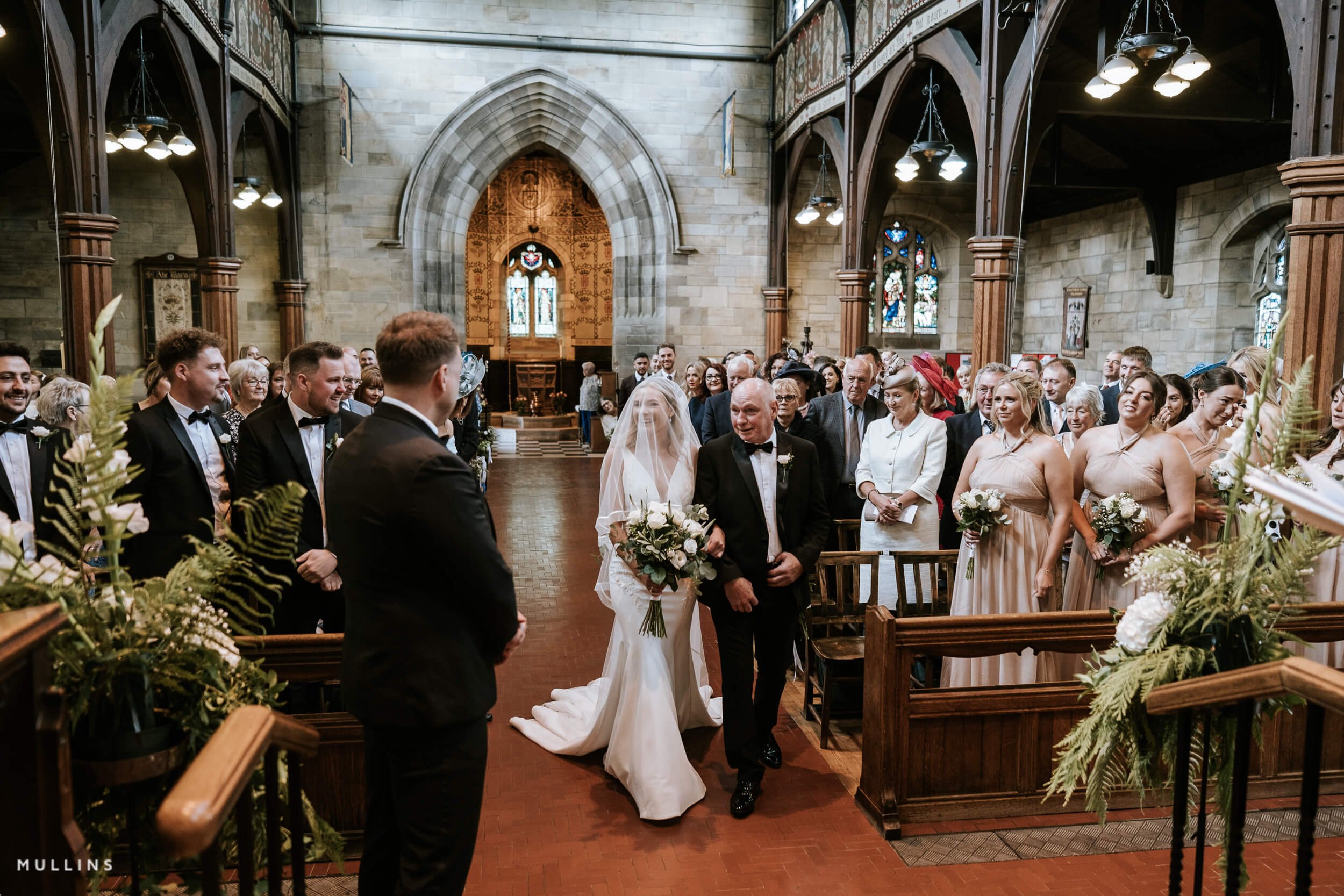 Bride walking down the aisle with her father during a wedding ceremony inside a church, surrounded by seated guests and stained glass windows.