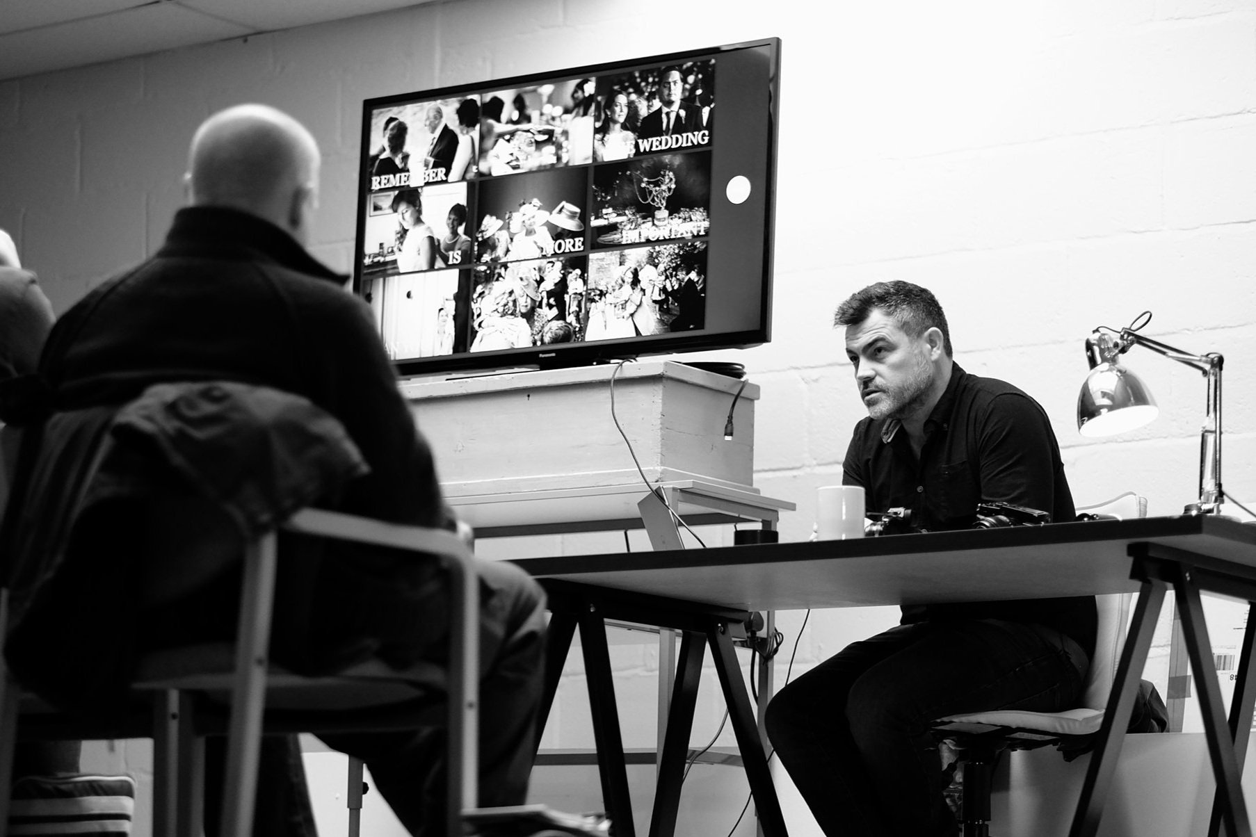 A black-and-white photo of a man with short hair sitting at a desk, facing a few people in a room. On the wall behind him is a large monitor displaying a collage of images, including wedding photos and people at an event.