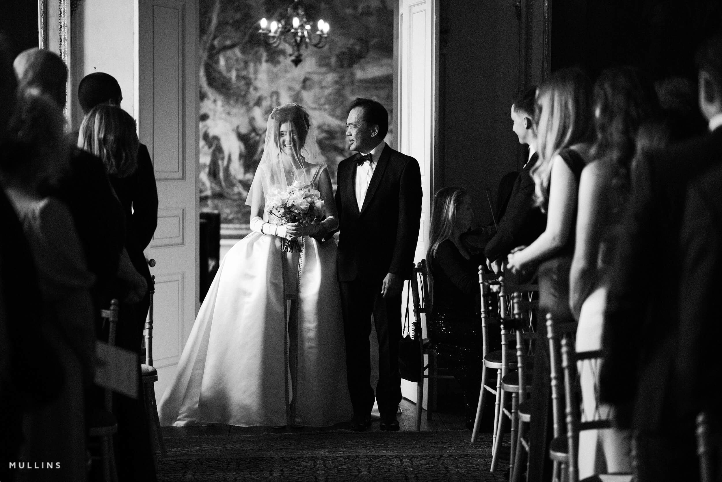 Black and white photo of a bride and an older man walking into a wedding ceremony, surrounded by seated and standing guests in an ornate room with a chandelier and artwork on the wall.