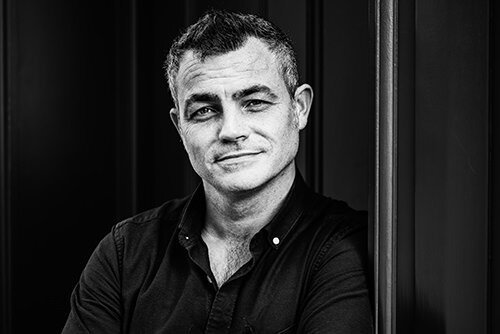 Black and white photo of a man with short dark hair, wearing a dark shirt, leaning against a dark wall, looking at the camera with a neutral expression.
