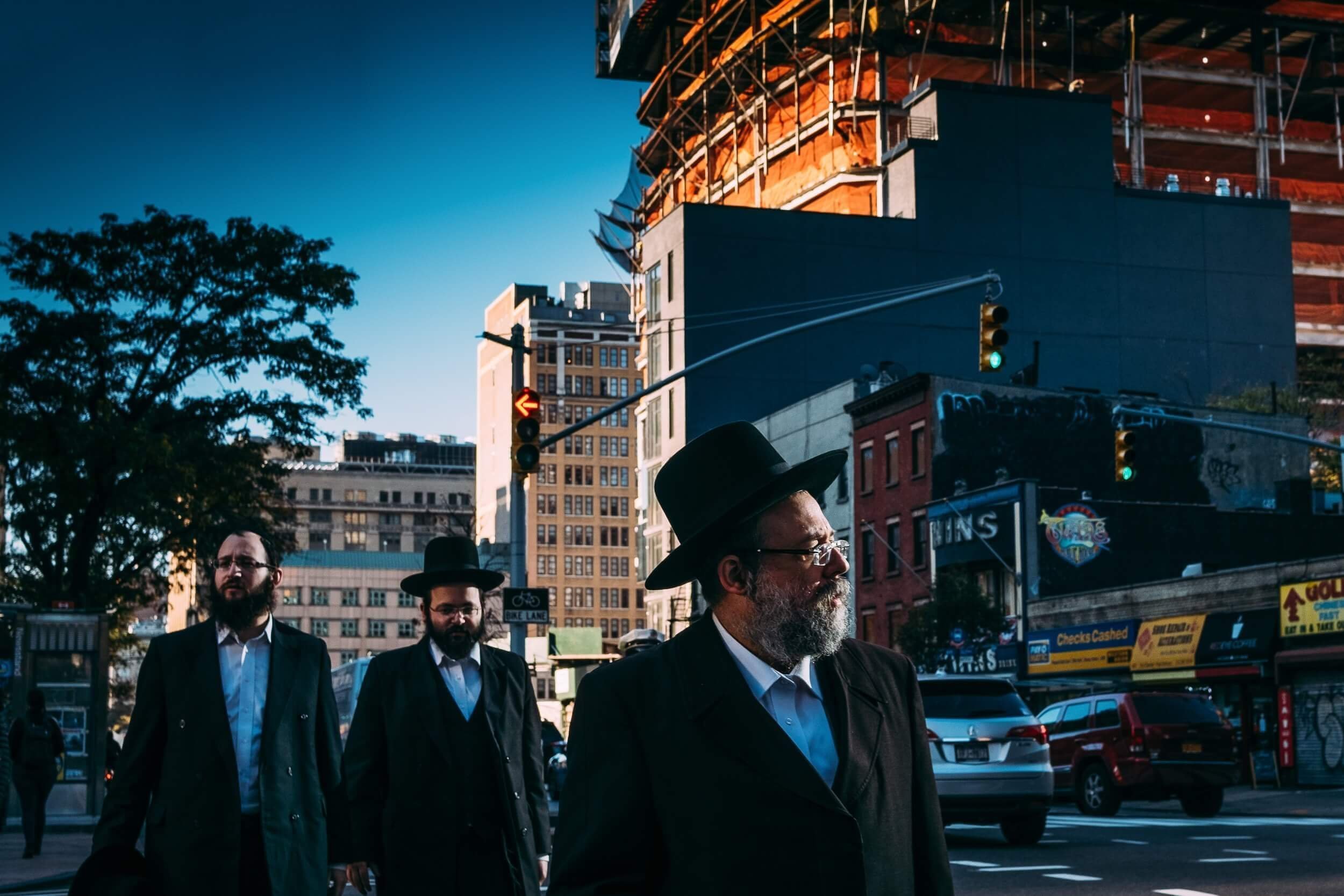 Three men with beards and glasses wearing black suits and black hats walking across a city street at dusk with tall buildings, traffic lights, and parked cars in the background.