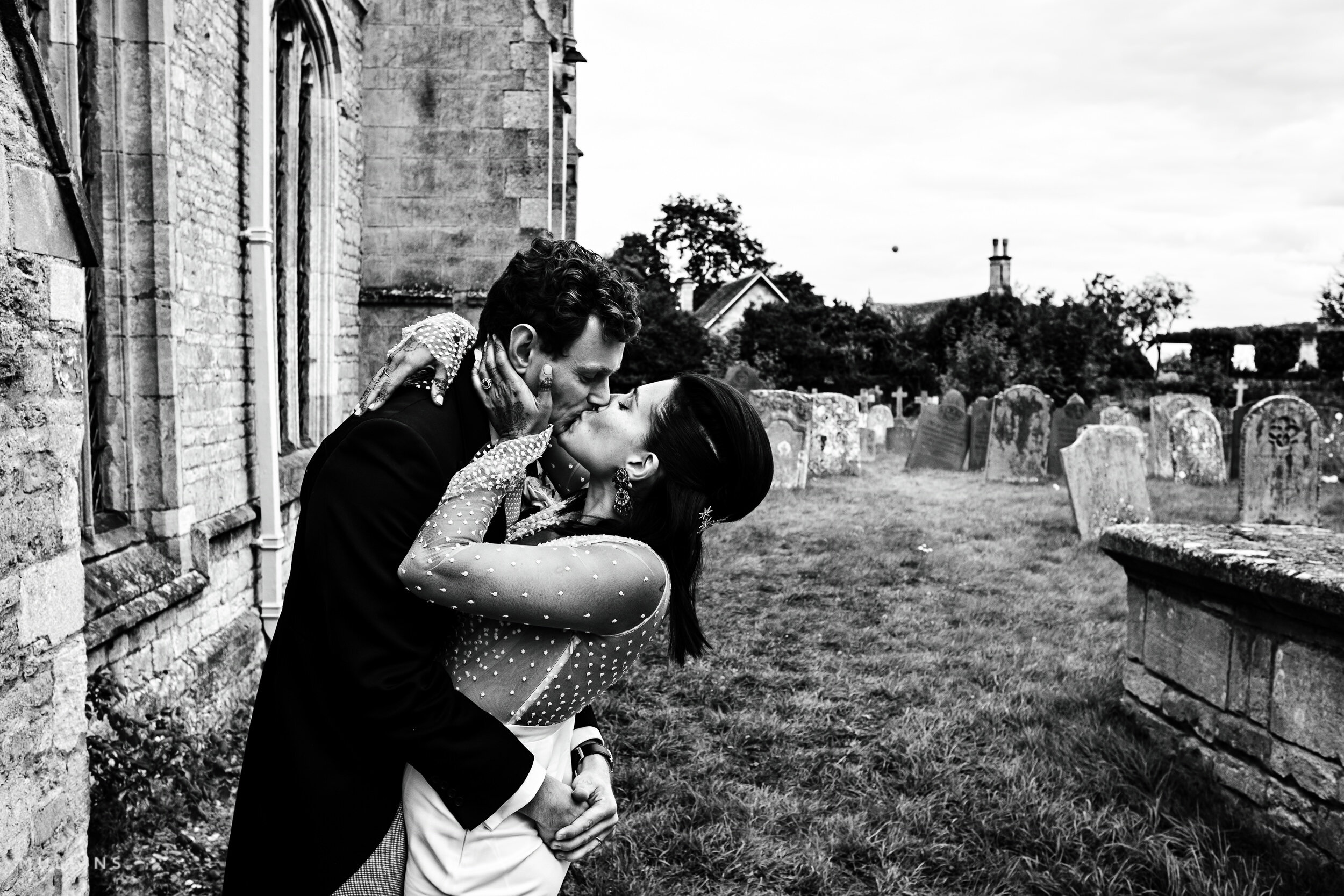 A couple in wedding attire sharing a kiss in a graveyard with headstones and trees in the background, black and white photo.